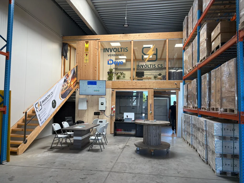 Lager, Showroom und Büro von Involtis in Landscha an der Mur: Galeriebüro mit den Schriftzügen INVOLTIS, Deye und Easyway, Treppe mit Involtis-Banner, Besprechungstisch, Wechselrichter an der Wand und Palettenregale mit verpackter Ware